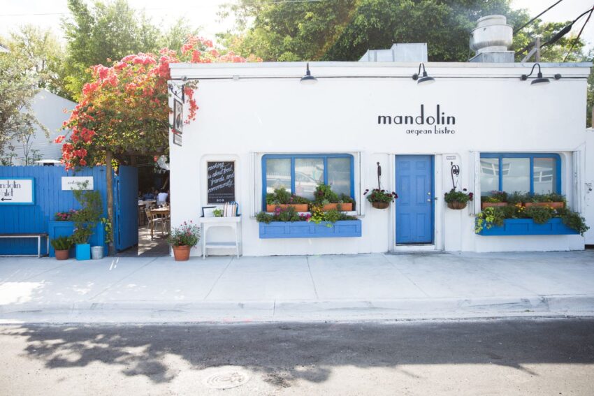 Exterior of Mandolin Aegean Bistro with blue doors, flowering plants, and a patio area under bright sunlight.