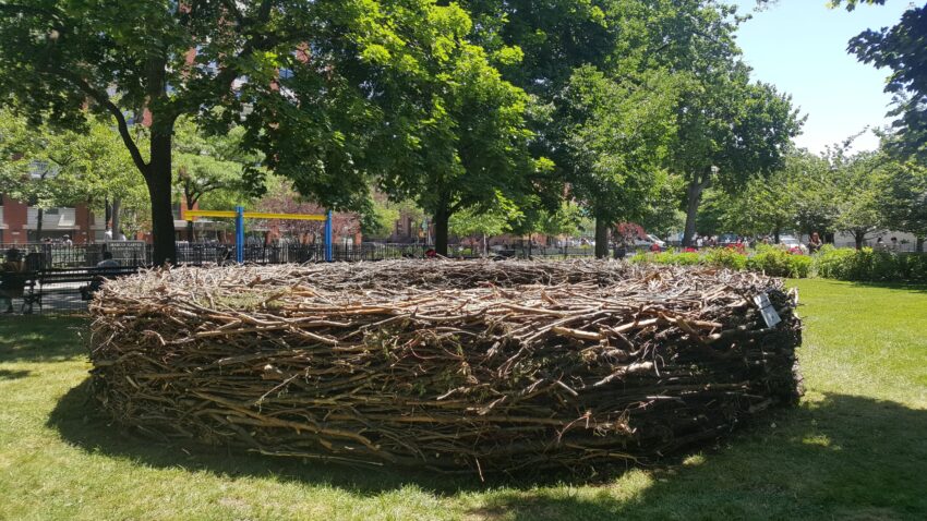 Large nest made of intertwined branches in a park, surrounded by lush trees and playground equipment in the background.