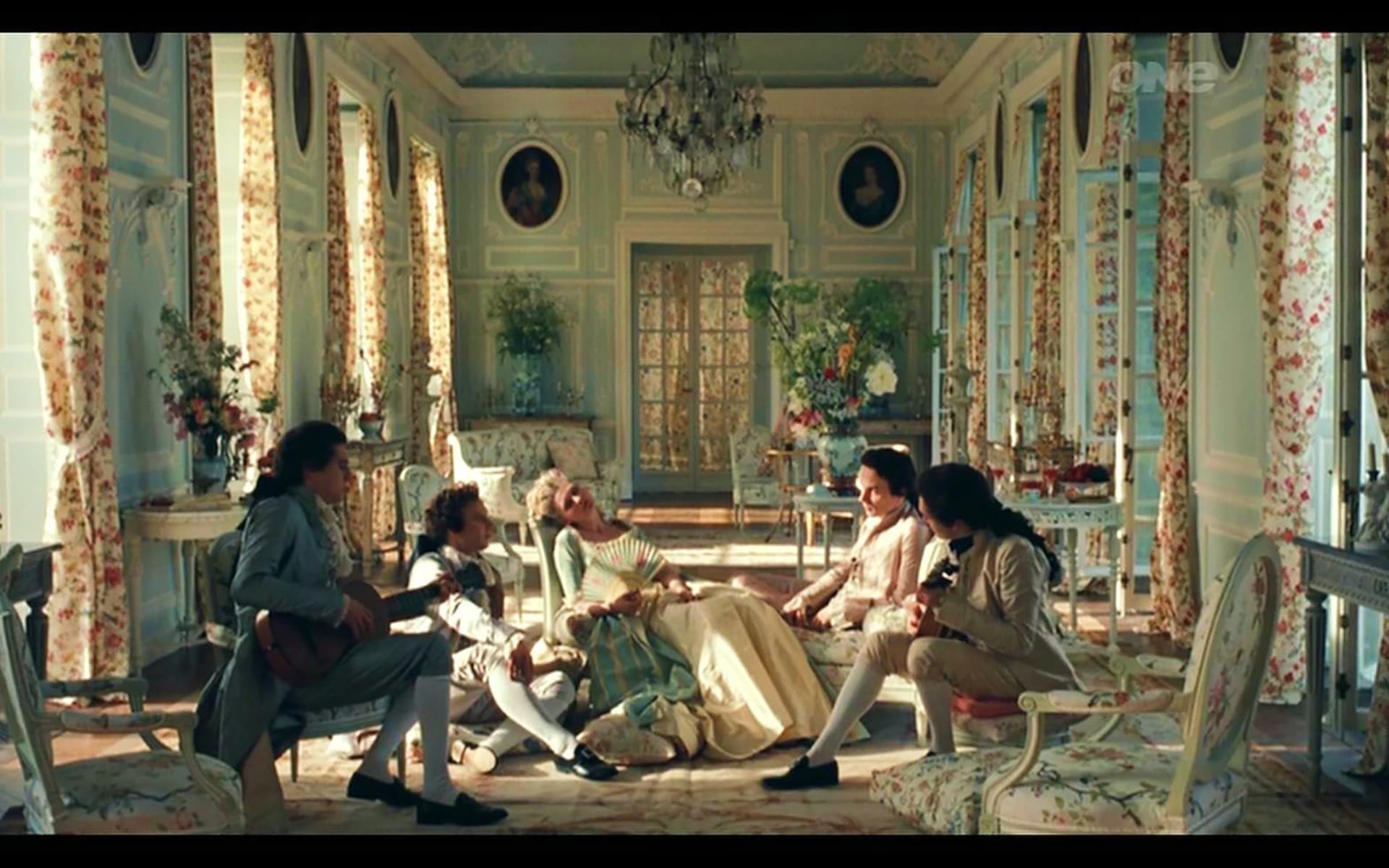 Historical figures in 18th-century attire relaxing in an ornate, floral-decorated room with large windows and chandeliers.