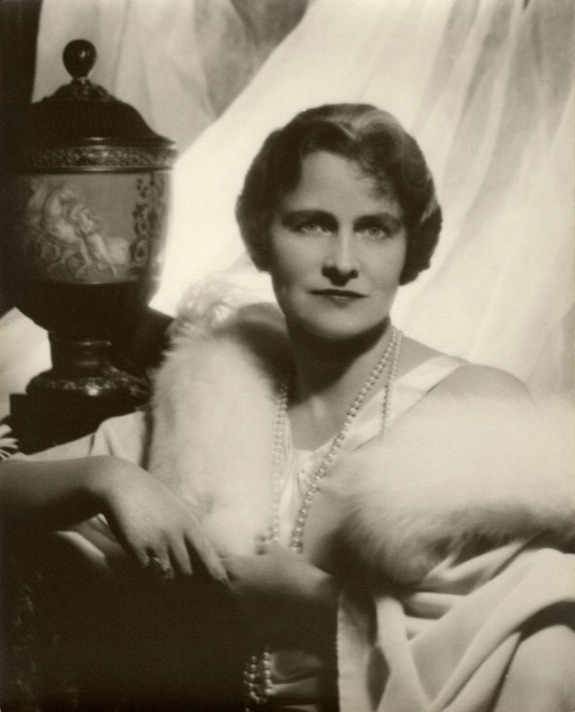 A person in vintage attire with pearls, seated near an ornate vase, against a backdrop of sheer curtains.