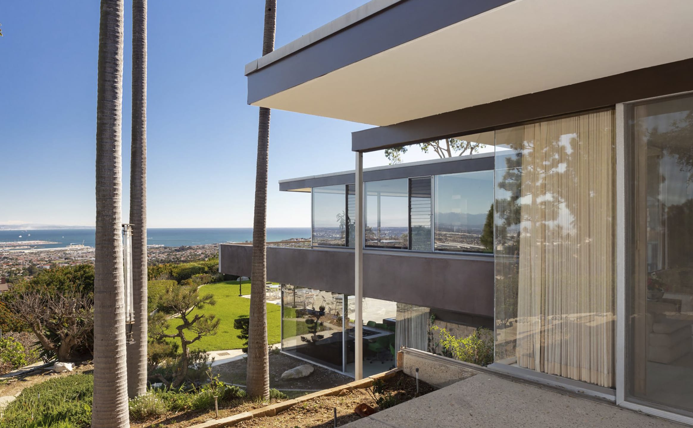 Modern glass-walled house with ocean view, surrounded by palm trees and a well-manicured lawn under a clear blue sky.