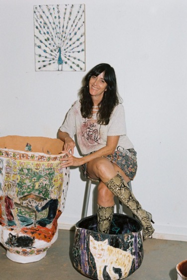Person sitting in a studio surrounded by large, decorative pottery and a peacock painting on the wall.
