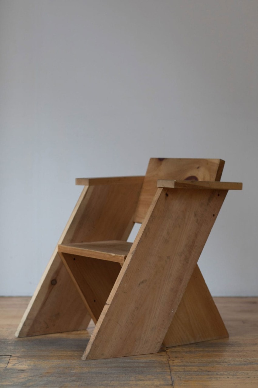 Wooden chair with angled legs and armrests on a wooden floor in a minimalist room setting.
