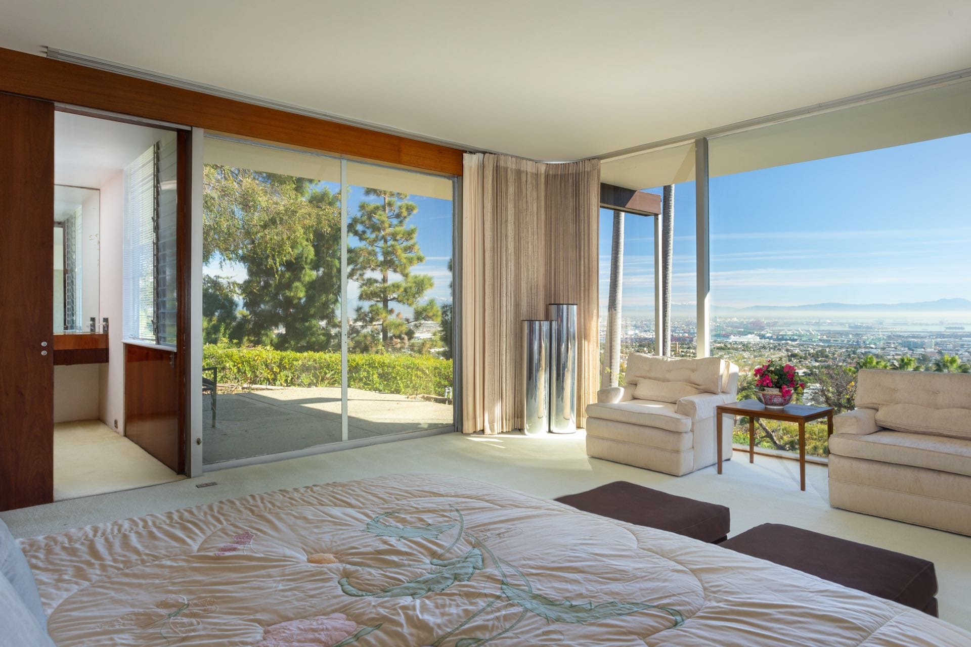 Spacious bedroom with large windows offering a view of the city, featuring modern furniture and a floral-patterned bedspread.