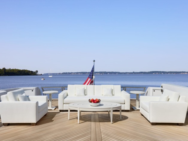 Luxury yacht deck with white sofas and table, overlooking calm water and forested shoreline, American flag visible in distance.
