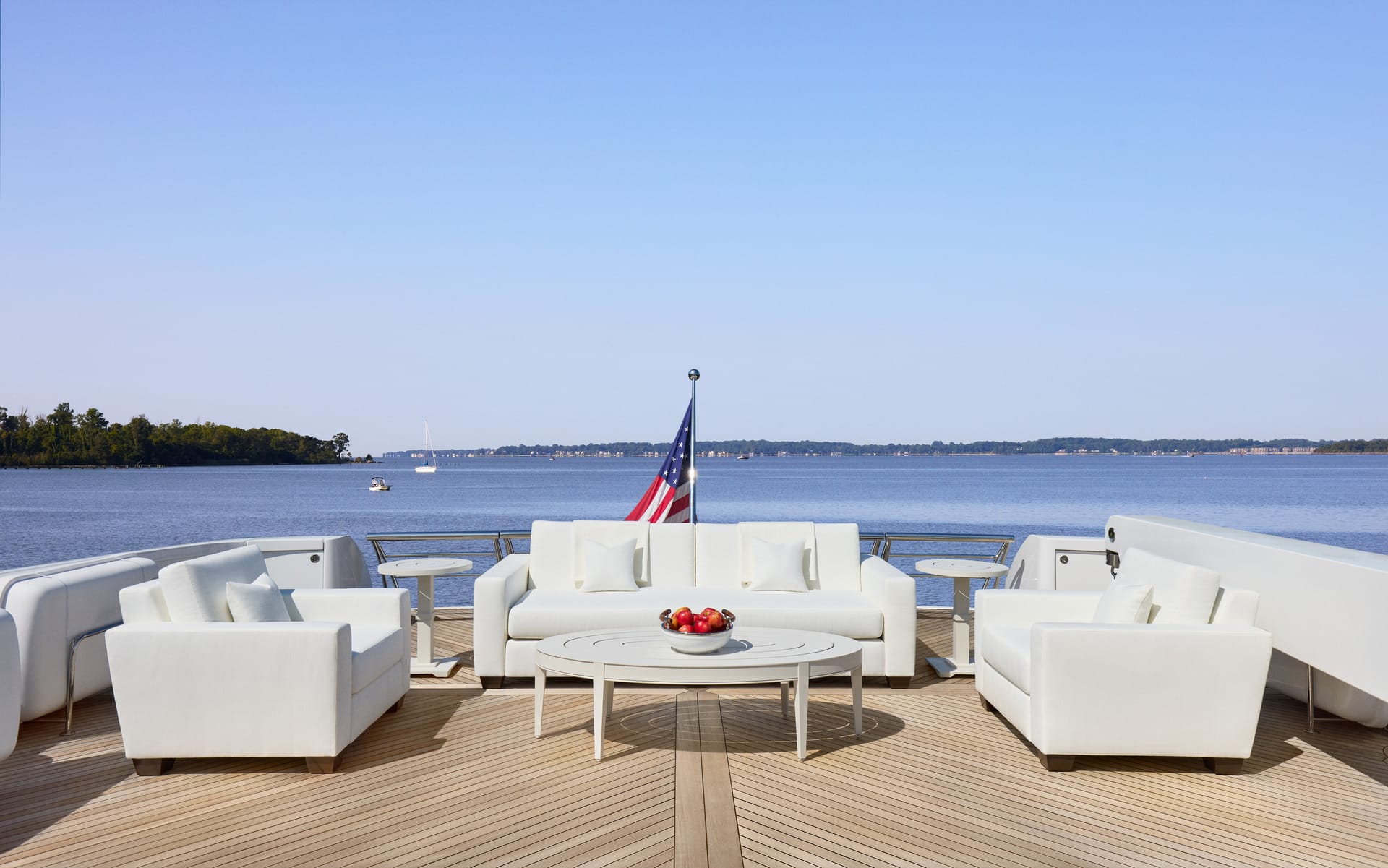 Luxury yacht deck with white sofas and table, overlooking calm water and forested shoreline, American flag visible in distance.