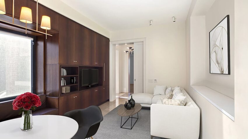 Modern living room with white sofa, round coffee table, and built-in wooden shelves. Large window with sunlight.