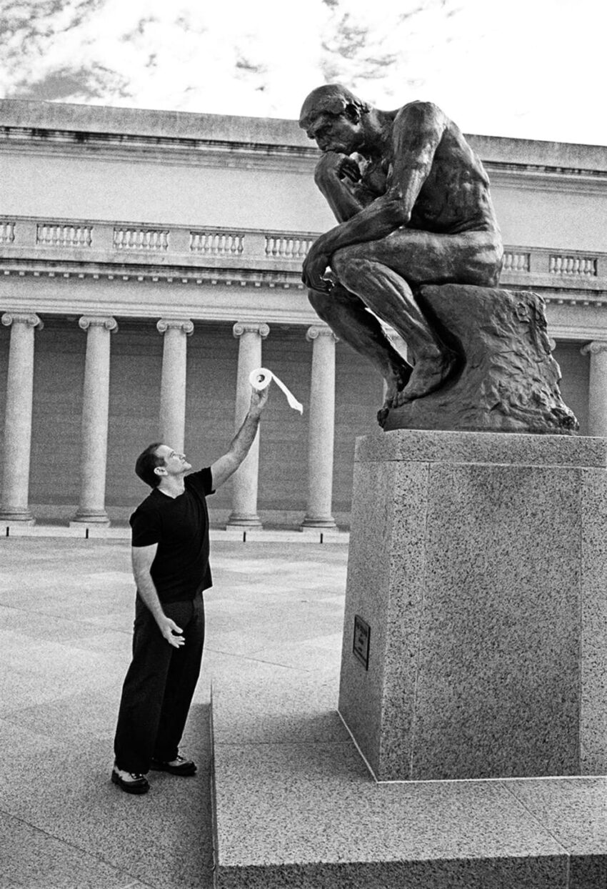 Man playfully reaching up with toilet paper towards a large sculpture outdoors.