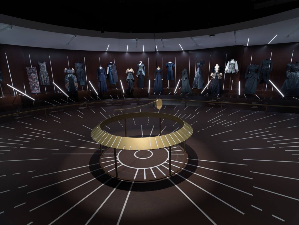 Fashion exhibition room with spotlighted dresses and a central circular display table in a dimly lit space.