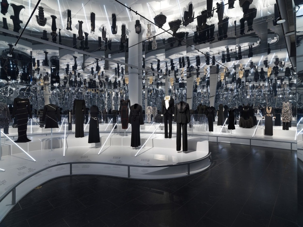 Fashion exhibit showcasing black garments in a mirrored room with sleek displays and reflective surfaces.