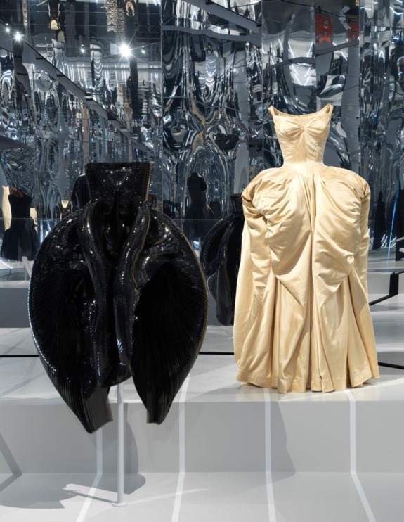 Two sculptural dresses on display; one black and abstract, the other cream with dramatic folds, in a mirrored exhibition space.