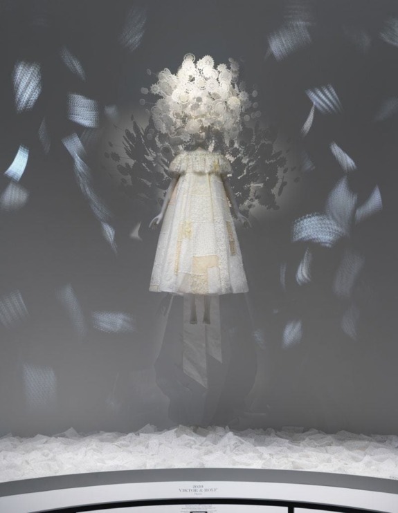 White floral dress on display against a blurred, abstract background in a museum setting.