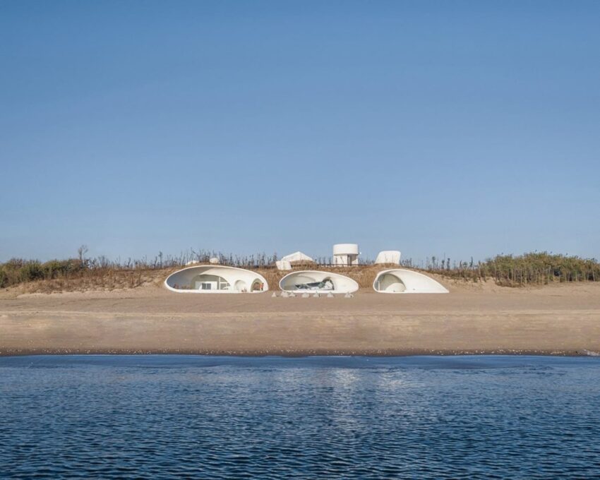 Modern beach house with unique arches and circular windows, situated on a sandy shore with clear blue sky and calm water.