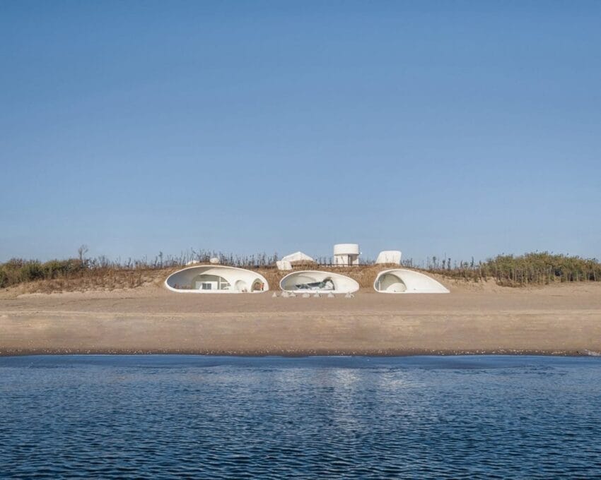 Modern beach house with unique arches and circular windows, situated on a sandy shore with clear blue sky and calm water.