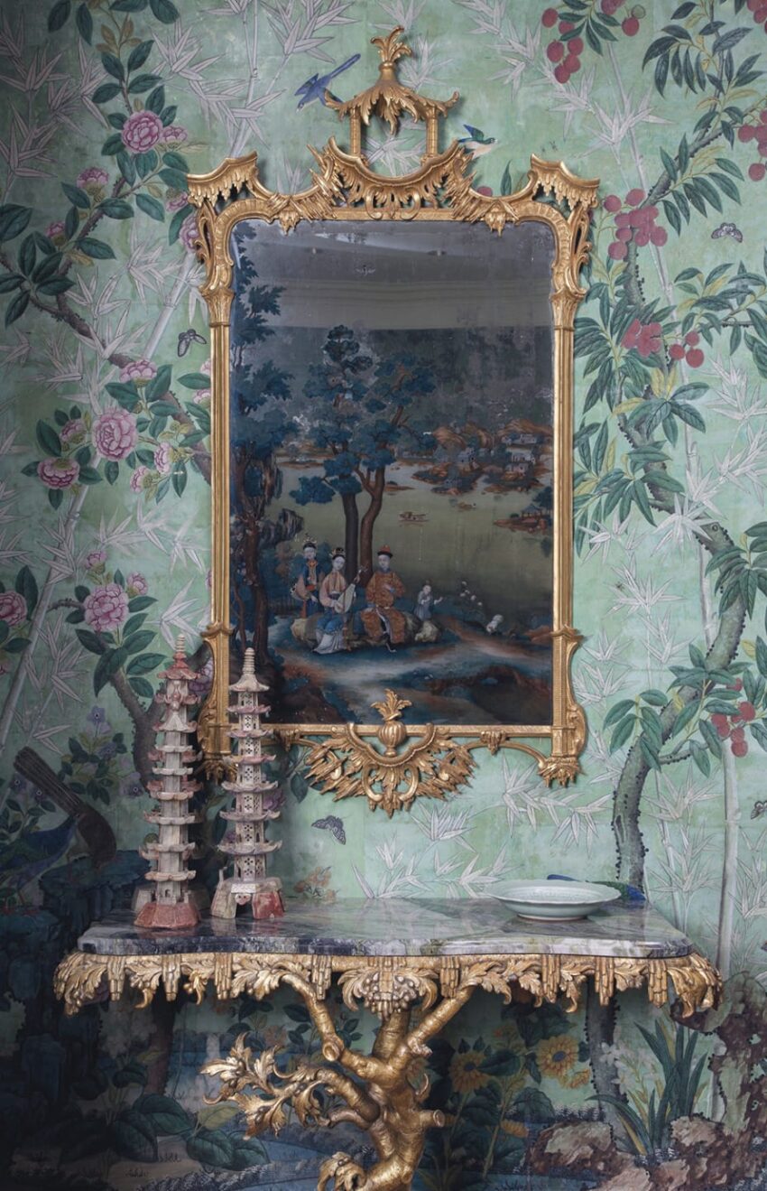 Ornate gold-framed mirror on a floral wallpapered wall, with a decorative table and miniature pagoda sculptures below.