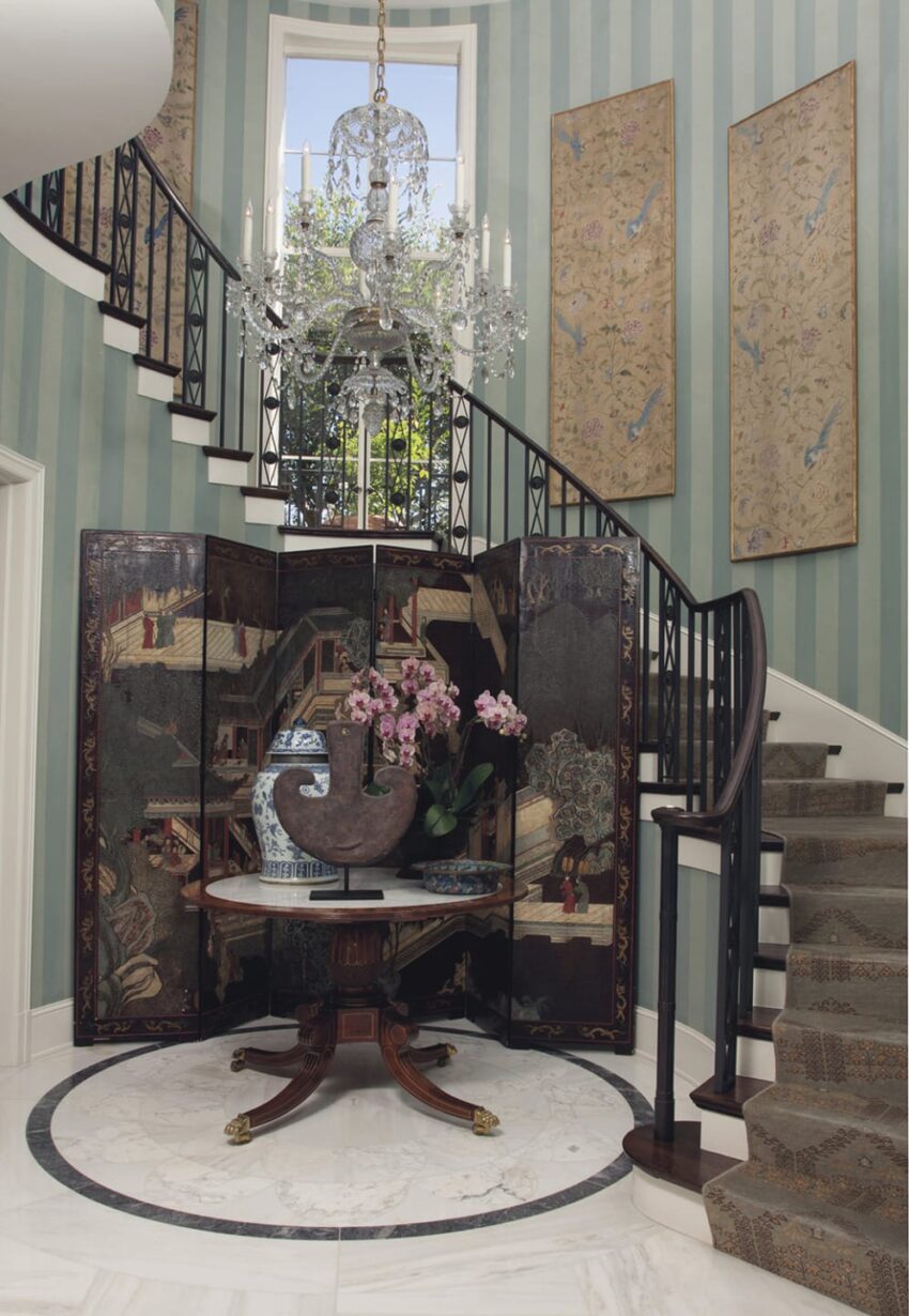 Elegant foyer with striped walls, crystal chandelier, decorative screen, round table, vase with pink orchids, curved staircase.