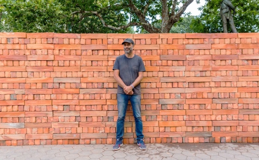 Man standing in front of a wall made of stacked orange bricks surrounded by trees in a sunlit outdoor setting.