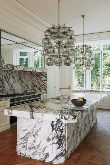 Luxurious kitchen with marble island, gold fixtures, and large glass chandeliers against a backdrop of large windows and greenery.