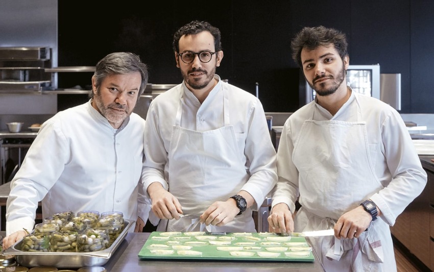 Three chefs in white uniforms preparing dishes in a professional kitchen.