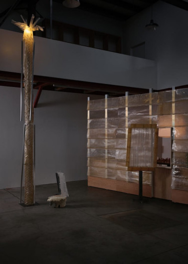 Art installation featuring tall illuminated structure, a wooden panel with a square cutout, and an irregularly shaped seat.