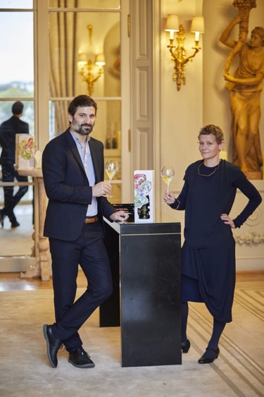 Two people standing by a pedestal holding wine glasses in an elegantly decorated room with artwork on the walls.