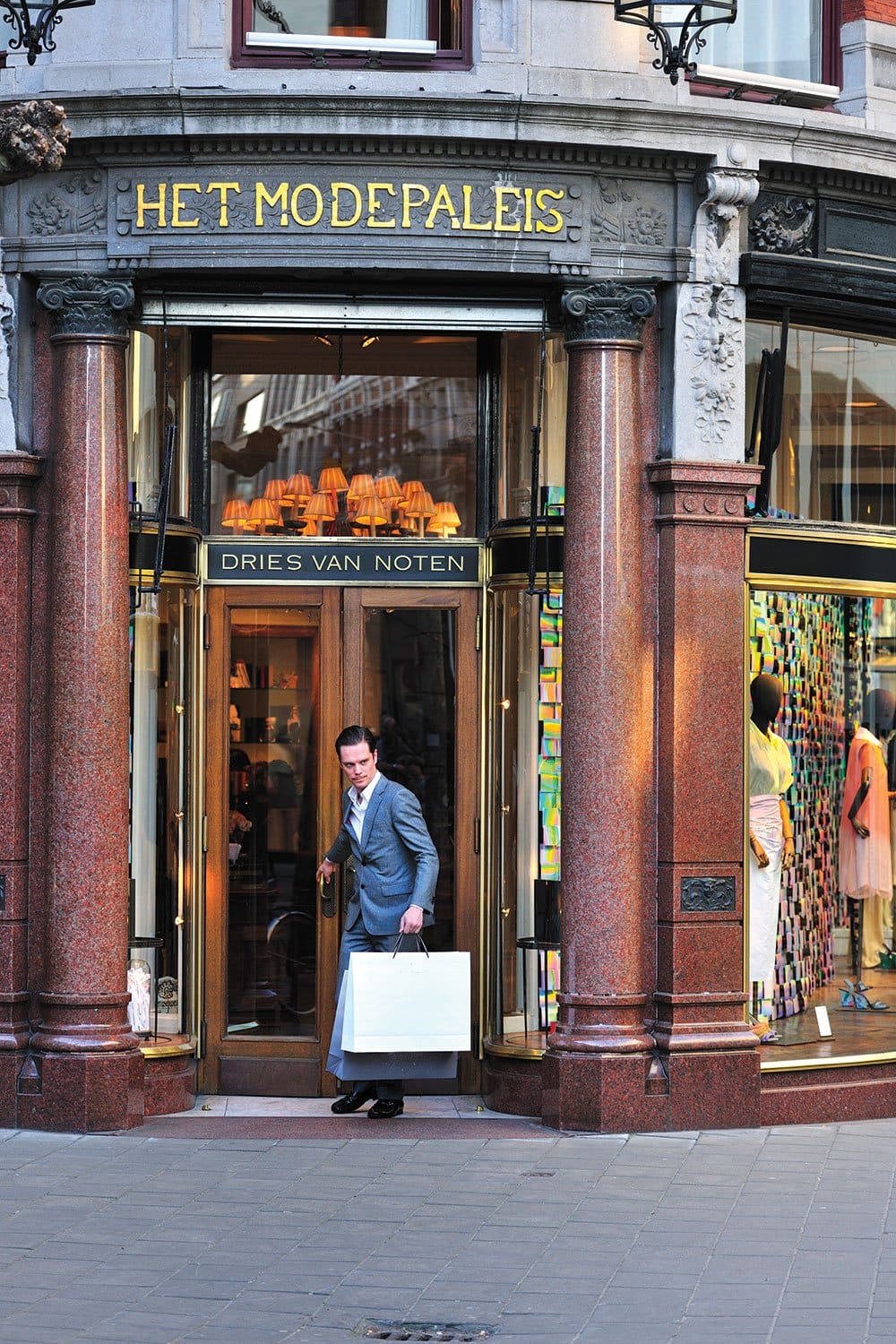 Man in suit exiting store with shopping bags; storefront features ornate columns and colorful displays inside.