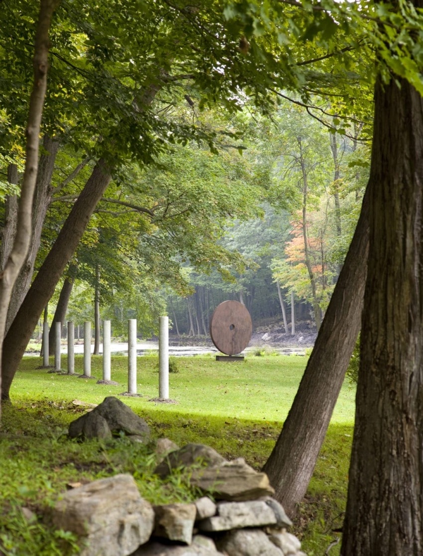 Art installation in a lush forest setting with trees, stone pillars, and a large circular sculpture on grassy ground.