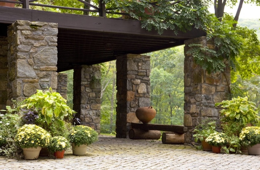 Stone patio with large pots of various plants and flowers, surrounded by lush greenery and a wooden overhang.