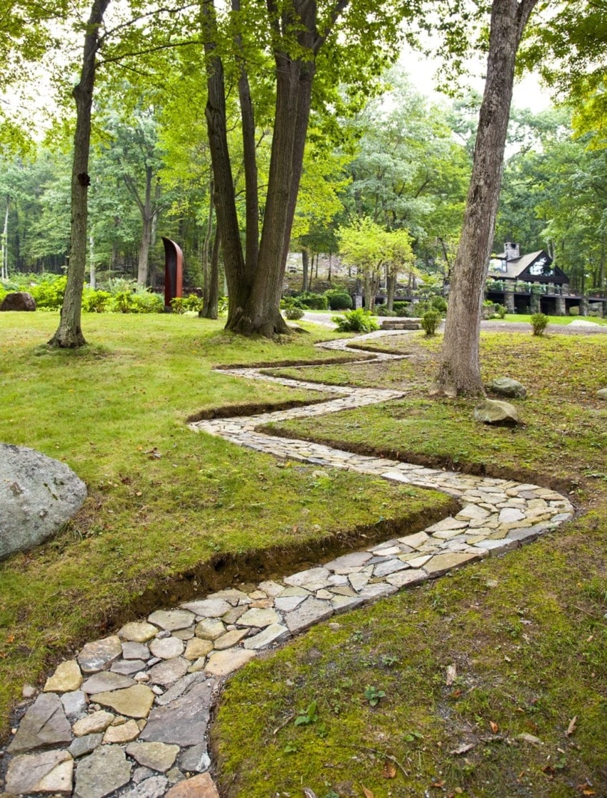 Winding stone path through a lush green forest with tall trees and grassy surroundings on a bright day.
