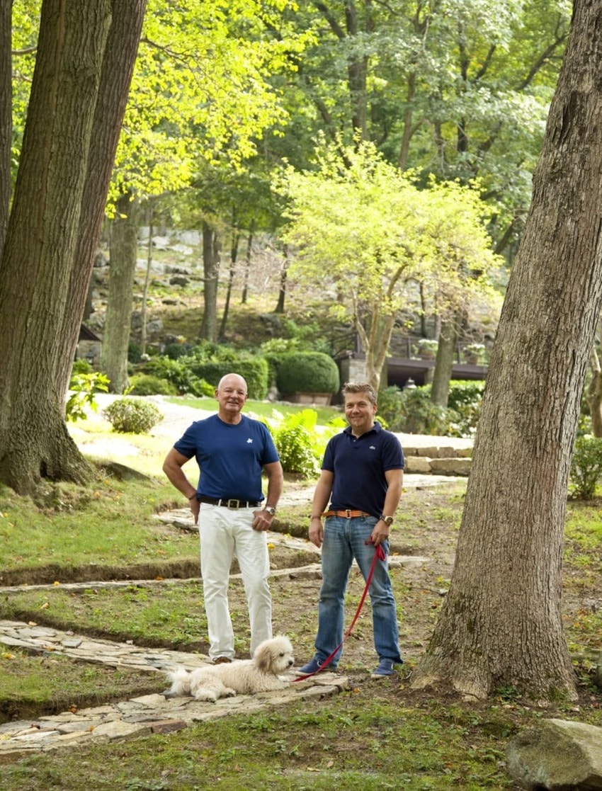 Two people standing on a wooded path with a dog on a leash, surrounded by trees and greenery.