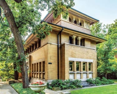 Two-story brick house with ornate windows surrounded by lush greenery and trees.