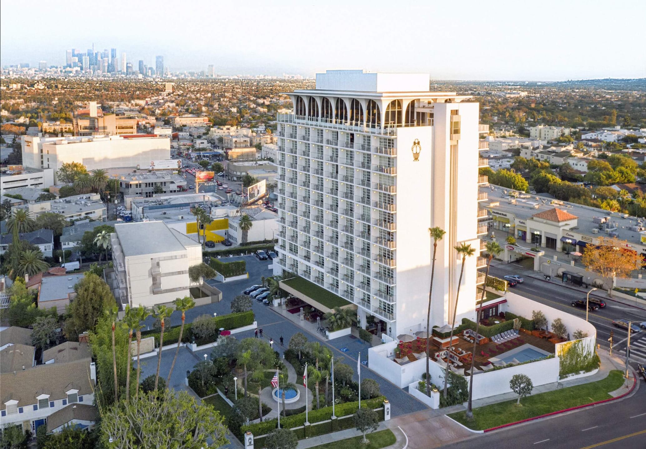 Aerial view of a tall white hotel surrounded by urban landscape with downtown skyline in the distance.