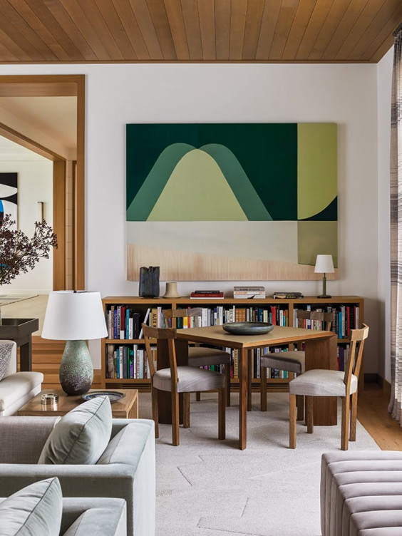 Modern living room with abstract artwork, wooden furniture, bookshelves filled with books, and soft gray couches.