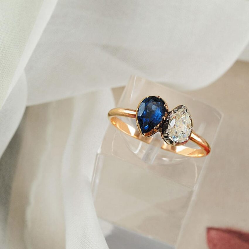 Gold ring with a pear-shaped blue sapphire and a pear-shaped diamond on a light fabric background.