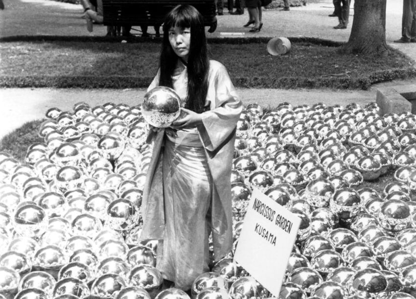 Artist standing among reflective spheres in a garden display, holding a sphere, with a sign reading "Narcissus Garden Kusama".