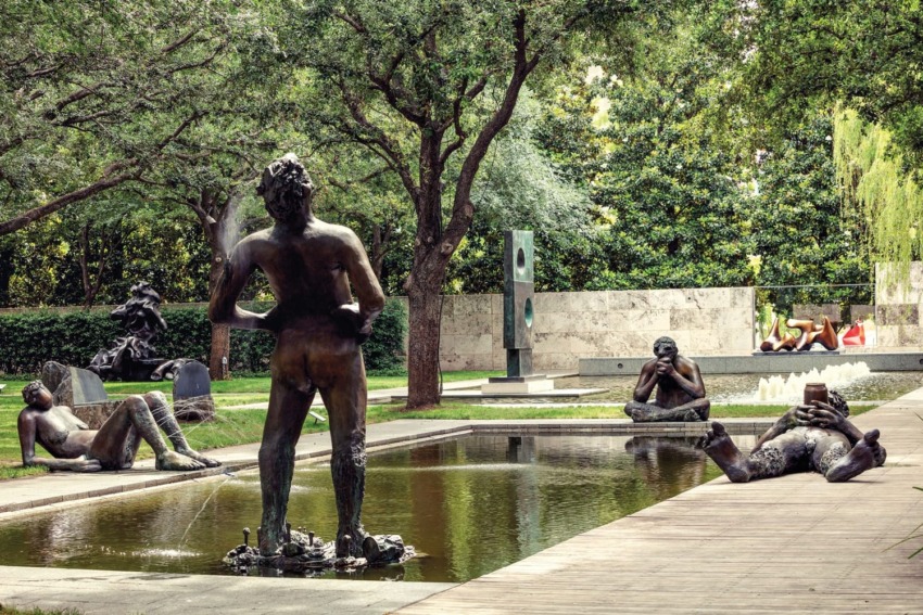 Sculpture garden featuring statues around a reflective pond, surrounded by trees and greenery, with fountains in the background.