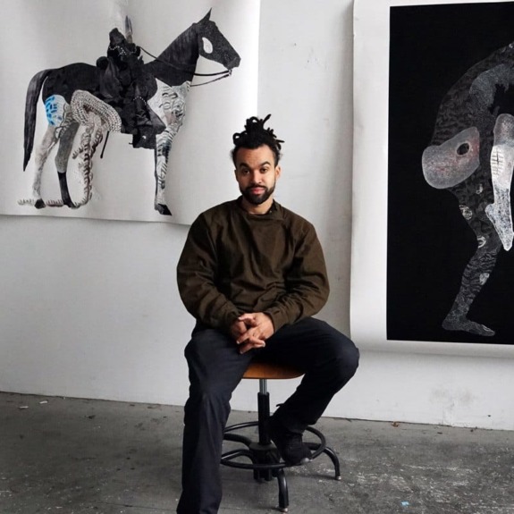 Artist seated on a stool in an art studio with two large artworks of horses displayed on the walls in the background.