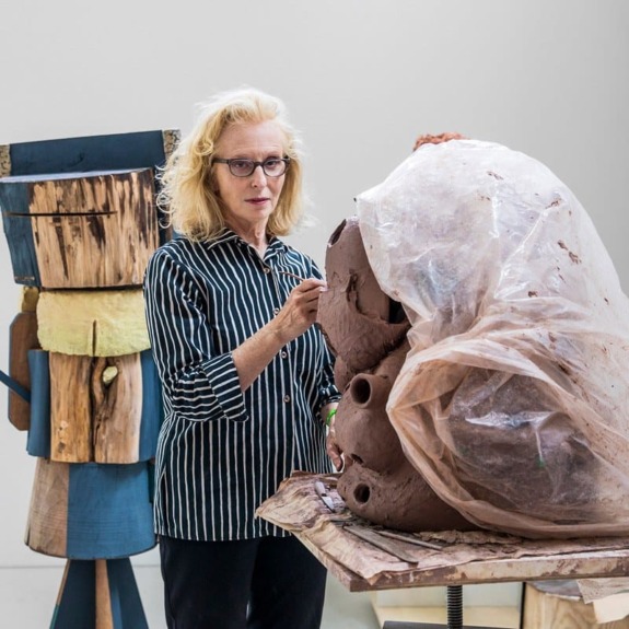 Artist sculpting clay, surrounded by abstract wooden sculptures in a studio.