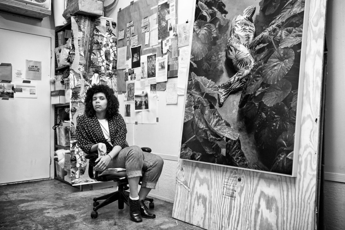 Artist seated in studio with large monochrome artwork, surrounded by various creative materials and pinned notes on the walls.