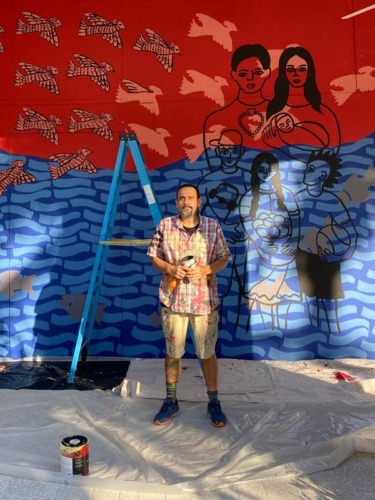 Artist standing in front of colorful mural with birds, people, and waves, holding paint supplies on a plastic-covered floor.