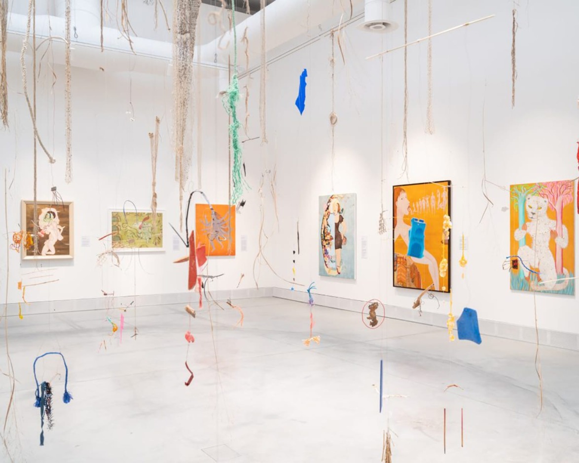Contemporary art exhibit with colorful paintings on white walls and hanging mixed media installations in a bright gallery space.
