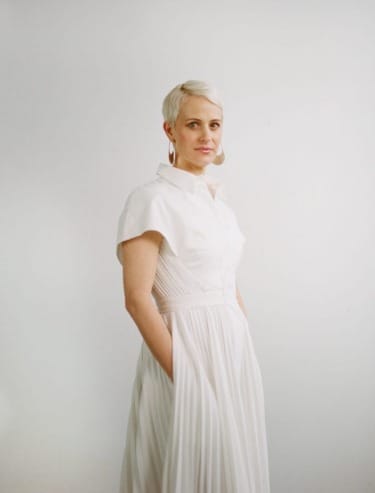 Person with short blonde hair wearing a white dress, standing against a plain background, looking confidently at the camera.