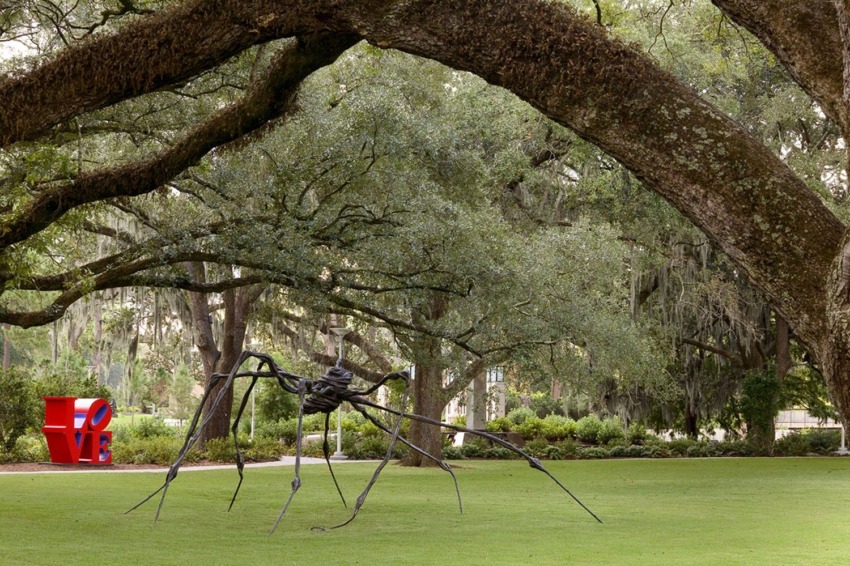 Large spider sculpture on a grassy lawn with trees and a red LOVE sculpture in the background