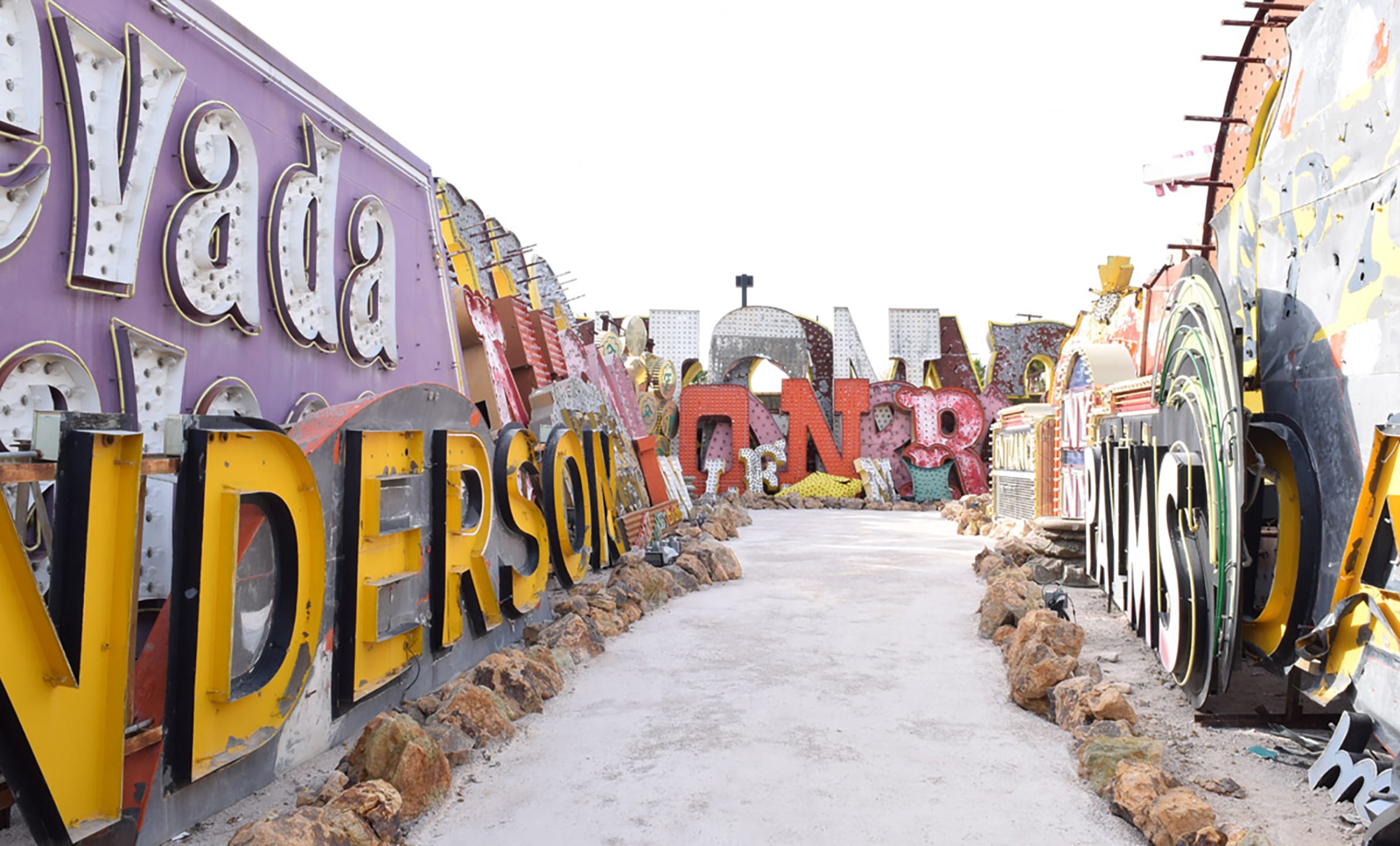 Pathway through vintage neon signs displayed outdoors against a bright sky.
