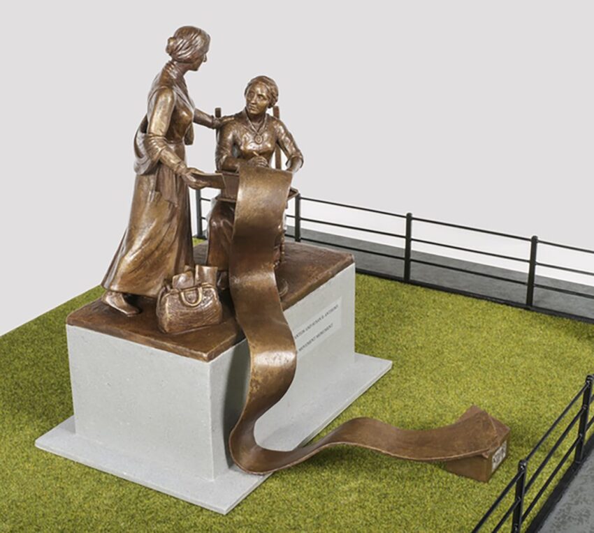 Statue of two women with scroll, one standing with a bag, the other seated, on a grassy base with a railing background.