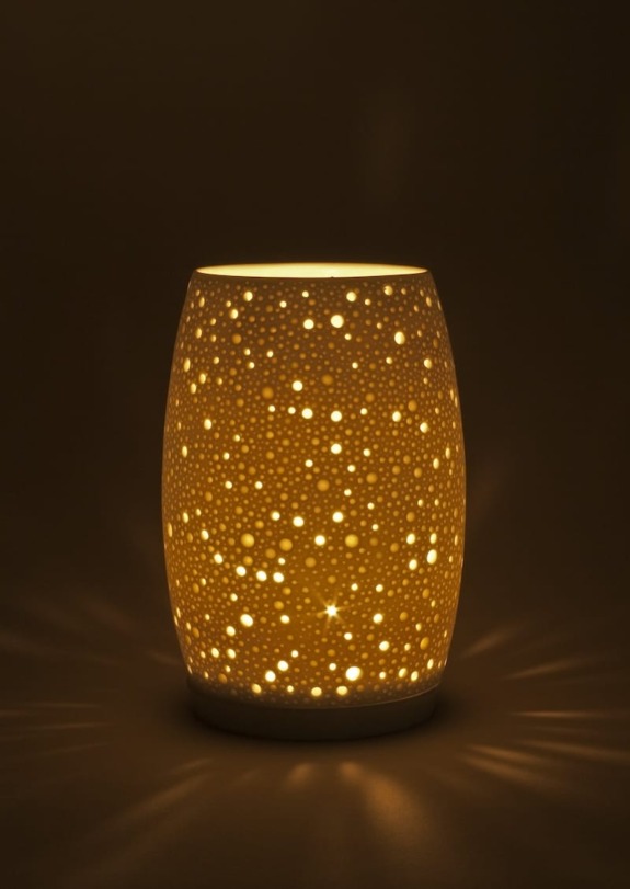 A ceramic lamp with star and dot cutouts glows warmly in a dark room, casting patterned light on surrounding surfaces.