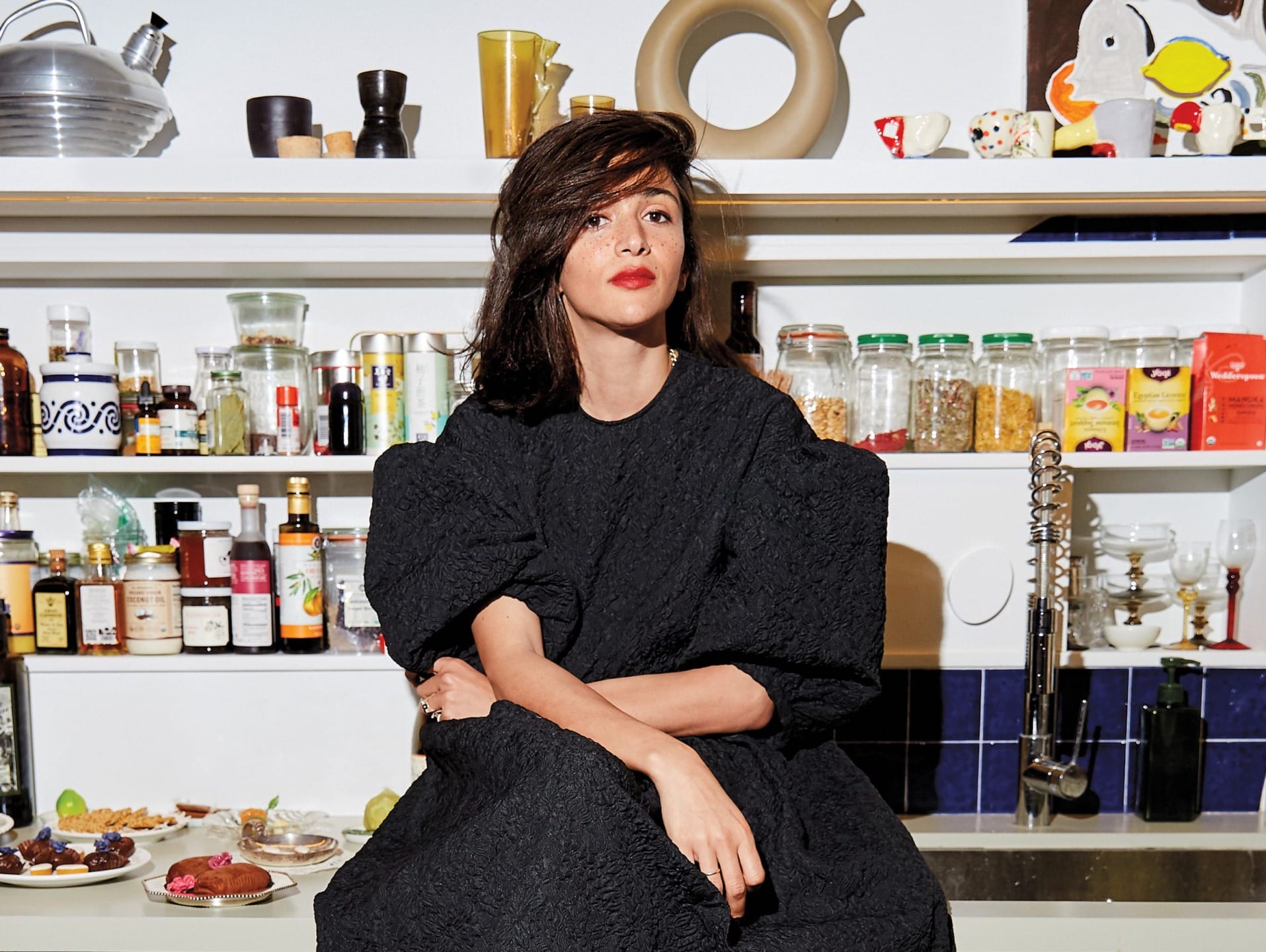 Person in a black dress sitting on a kitchen counter with shelves filled with jars and kitchen items in the background.