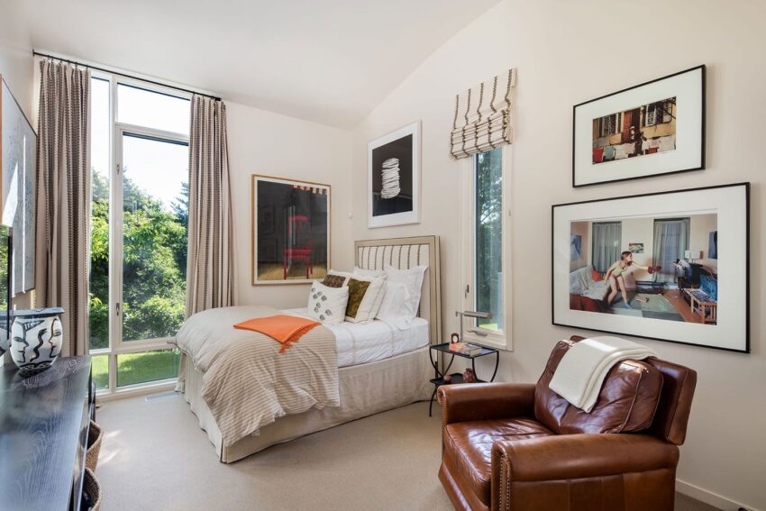 Cozy bedroom with large window, stylish decor, leather armchair, and framed artwork on cream-colored walls.