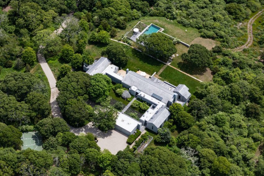 Aerial view of a large white house surrounded by trees, with a tennis court and pool in the backyard.