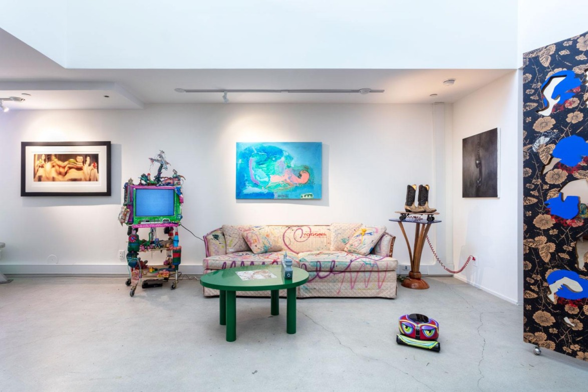 Eclectic art gallery with paintings on walls, a decorated TV, a patterned couch, a green table, and various artistic objects.